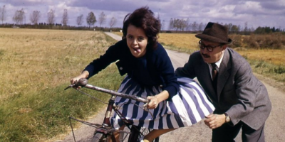 chica en una bicicleta sacando la lengua seguida por un hombre preocupado por que no se caiga